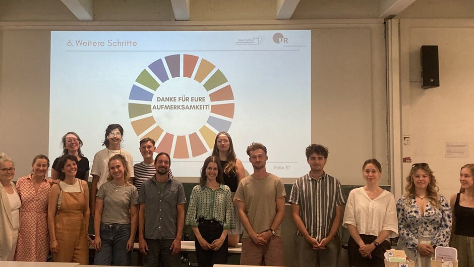Gruppenfoto von 15 Personen in einem Hörsaal der Universität Regensburg vor einer projizierten Präsentationsfolie zum Thema Nachhaltige Entwicklung. Auf der Folie ist ein kreisförmiges Symbol der Nachhaltigkeitsziele (SDGs) mit dem Schriftzug „Danke für eure Aufmerksamkeit!“ zu sehen. (öffnet Vergrößerung des Bildes)