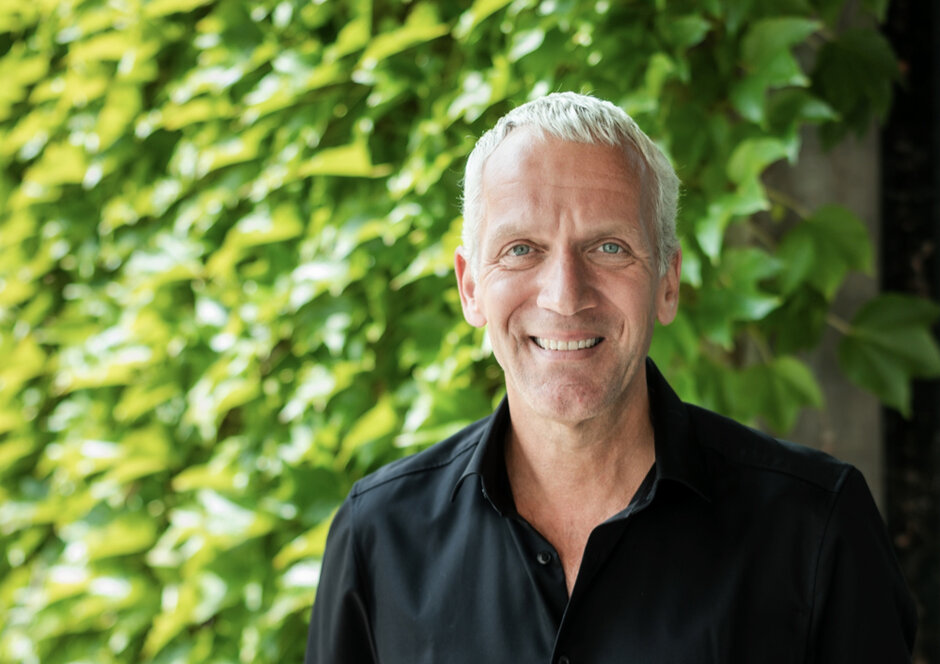Ein Mann mit grauen Haaren und schwarzem Hemd blickt lächelnd in Richtung des Betrachters. Im Hintergrund ist verschwommen eine mit einer grünen Kletterpflanze bewachsene Wand zu erkennen.