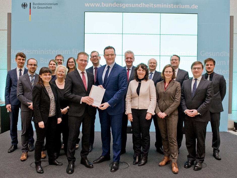 Das Bild zeigt die etwa 15 Mitglieder der Kommission mit Jens Spahn, der den Abschlussbericht in den Händen hält, auf einer Bühne mit dem Logo des Bundesgesundheitsministeriums. Bildquelle: BMG/Schinkel (öffnet Vergrößerung des Bildes)