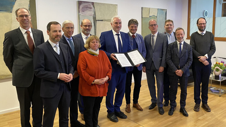 11 Personen in formeller Kleidung haben sich für ein Gruppenfoto aufgestellt. In der Mitte steht ein Mann, der eine aufgeschlagene Dokumentenmappe präsentiert. 