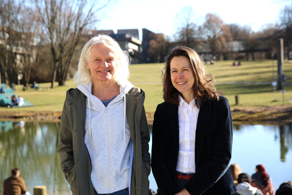 Zwei Frauen blicken lächelnd in die Kamera. Im Hintergrund sind ein Teich und eine weite Rasenfläche vor Hörsaalgebäuden zu sehen.