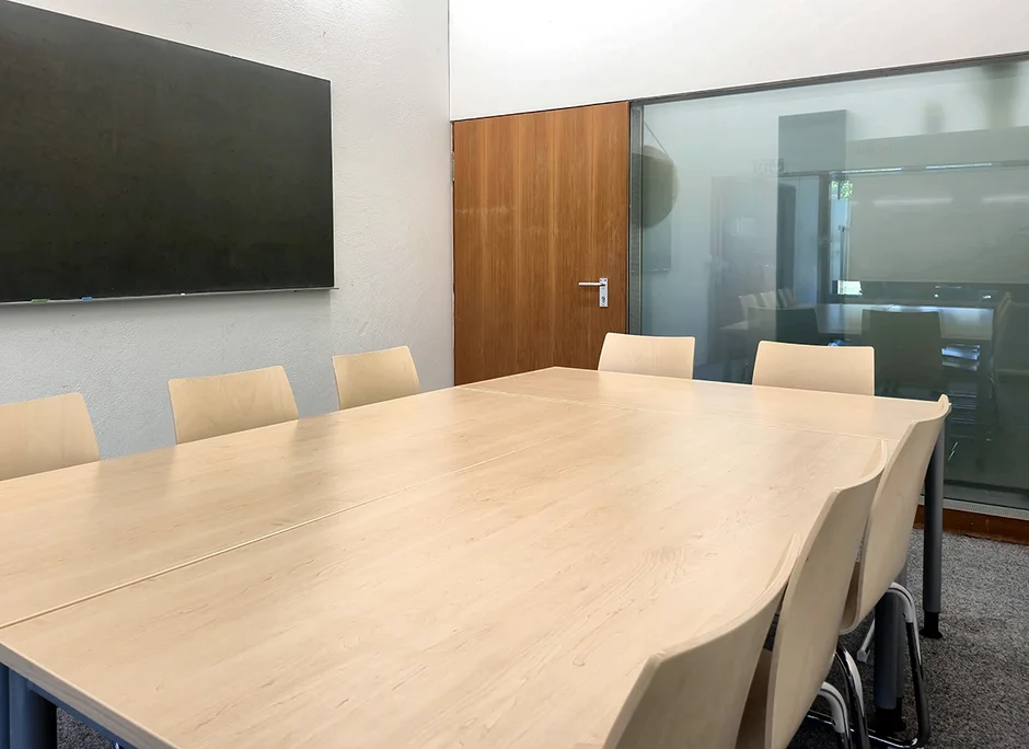 Gruppenarbeitsraum im Lesesaal Recht 1 mit großen Tischen und mehreren Holzstühlen. An der Wand hängt eine Tafel. Im Hintergrund befinden sich eine geschlossene Holztür sowie eine Glaswand. 