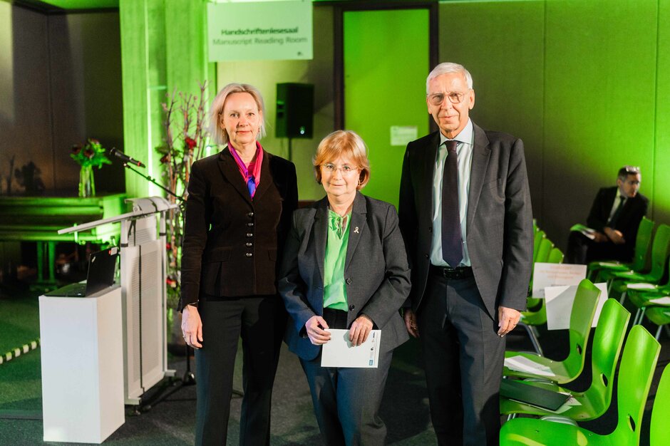 Zwei Frauen und ein Mann stehen nebeneinander und schauen in die Kamera. Im Hintergrund stehen ein Rednerpult und Stühle in der Universitätsbibliothek Regensburg.