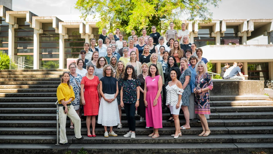 Gruppenfoto von rund 50 Personen auf einer breiten Außentreppe. Im Hintergrund steht ein großer, dicht belaubter Baum. Die Personen sind in mehreren Reihen angeordnet und tragen unterschiedlich farbige Kleidung. (öffnet Vergrößerung des Bildes)