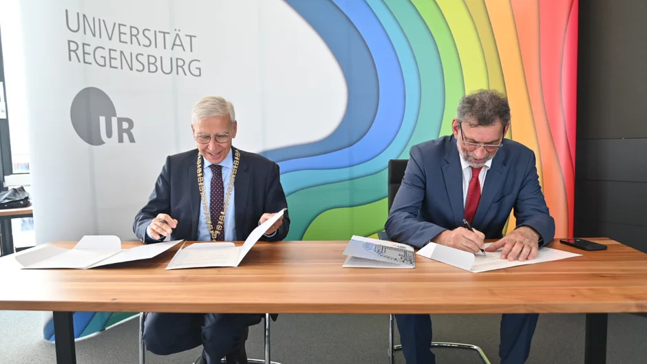 Zwei Männer in Anzügen und mit Krawatten sitzen an einem Konferenztisch und sind dabei, Papiere zu unterschreiben. Der eine Mann trägt eine Amtskette. Im Hintergrund ist ein vielfarbiger Aufsteller zu sehen, der den Schriftzug „Universität Regensburg“ trägt. (öffnet Vergrößerung des Bildes)