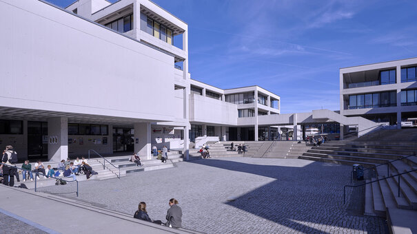 Gebäude ein brutalistischer Architektur der 1970er Jahre gruppieren sich um eine großzügige, autofreie Freifläche, die wie ein Amphitheater mit Sitzstufen angelegt ist. Studierende gehen oder sitzen einzeln oder in Gruppen auf dieser Fläche.