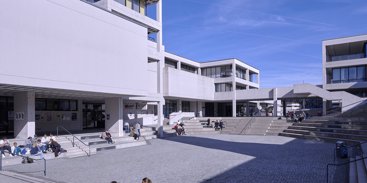 Gebäude ein brutalistischer Architektur der 1970er Jahre gruppieren sich um eine großzügige, autofreie Freifläche, die wie ein Amphitheater mit Sitzstufen angelegt ist. Studierende gehen oder sitzen einzeln oder in Gruppen auf dieser Fläche.