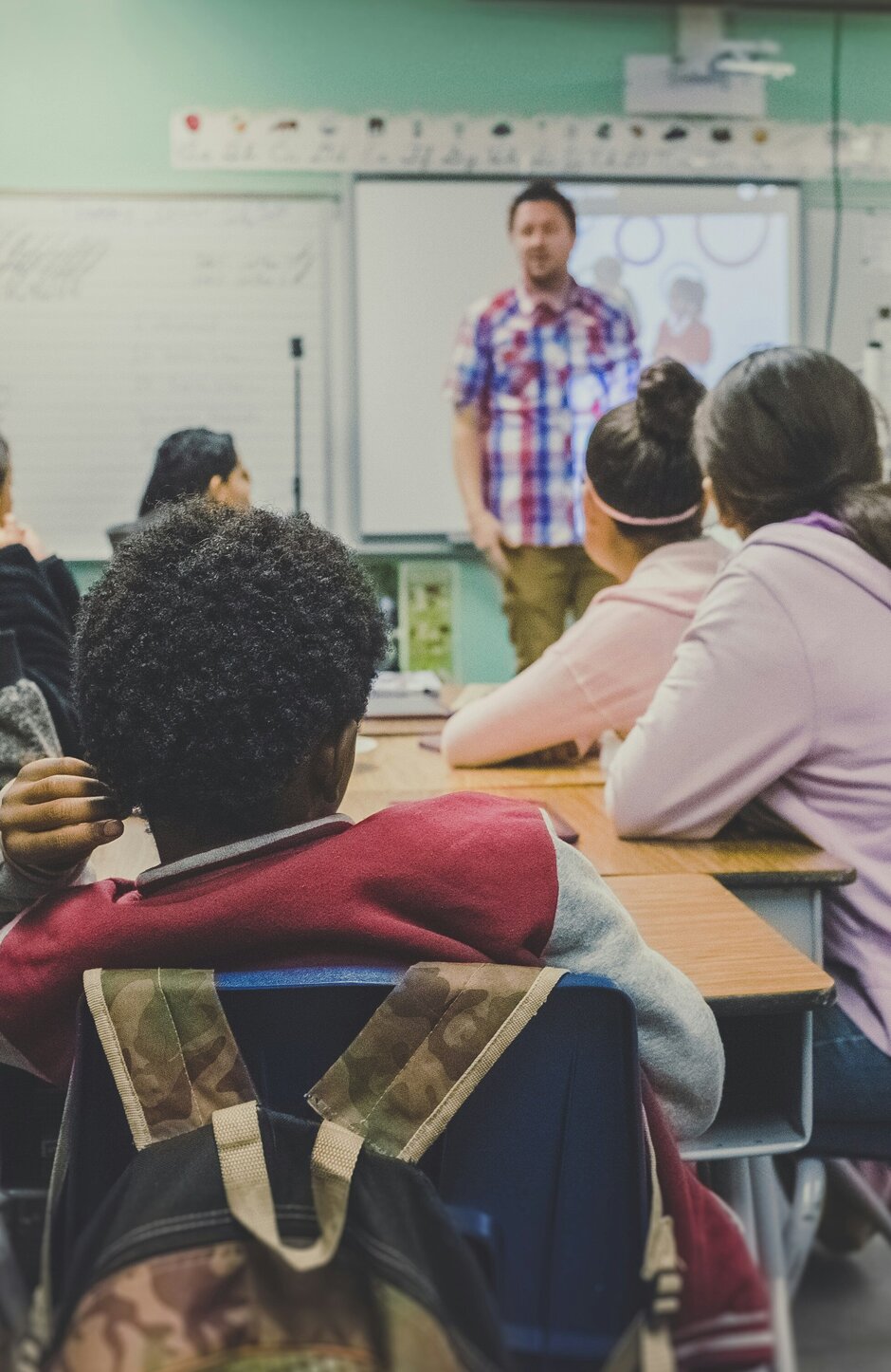 Klassenzimmer, die Schüler sitzen um Tische herum, vorn steht ein Lehrer