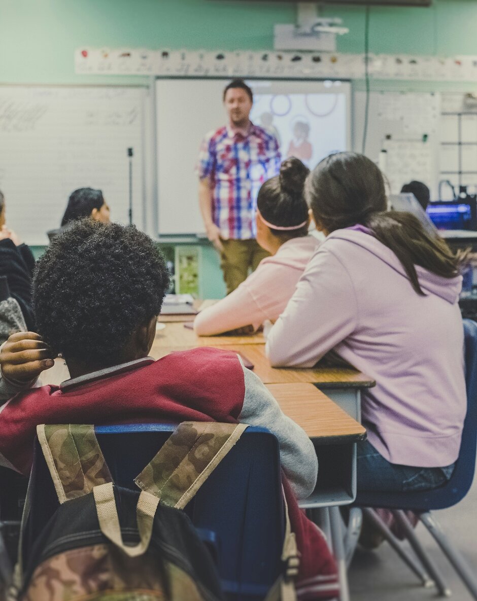 Klassenzimmer, die Schüler sitzen um Tische herum, vorn steht ein Lehrer