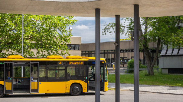 Ein gelber Linienbus mit dem Ziel "Universität" steht vor dem Sammelgebäude der Universität Regensburg