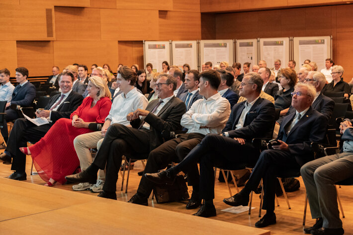 In einem Hörsaal sitzen viele Menschen im mittleren Alter in festlicher Garderobe auf mehreren hintereinanderstehenden Stuhlreihen. Die Atmosphäre wirkt gelöst. (öffnet Vergrößerung des Bildes)