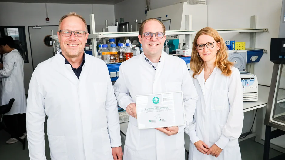 Drei Personen in Laborkitteln präsentieren in einem Labor eine Green-Lab-Urkunde der Universität Regensburg vor Laborgeräten und Arbeitsplätzen. (öffnet Vergrößerung des Bildes)