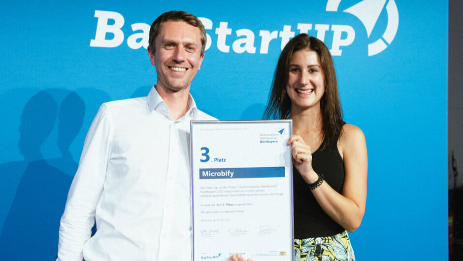 Ein Mann und eine Frau halten lächelnd eine Urkunde in die Kamera. Auf der Urkunde kann man den Schriftzug "3. Platz Microbify" erkennen. (öffnet Vergrößerung des Bildes)