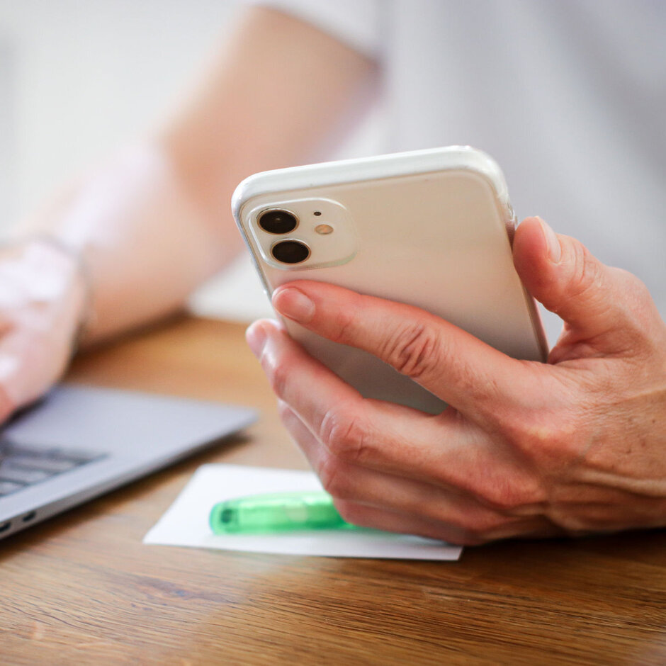 Eine Person hält ein Smartphone in der Hand und tippt etwas am Laptop.