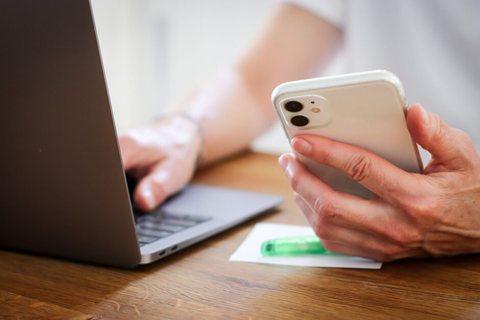 Eine Person hält ein Smartphone in der Hand und tippt etwas am Laptop.