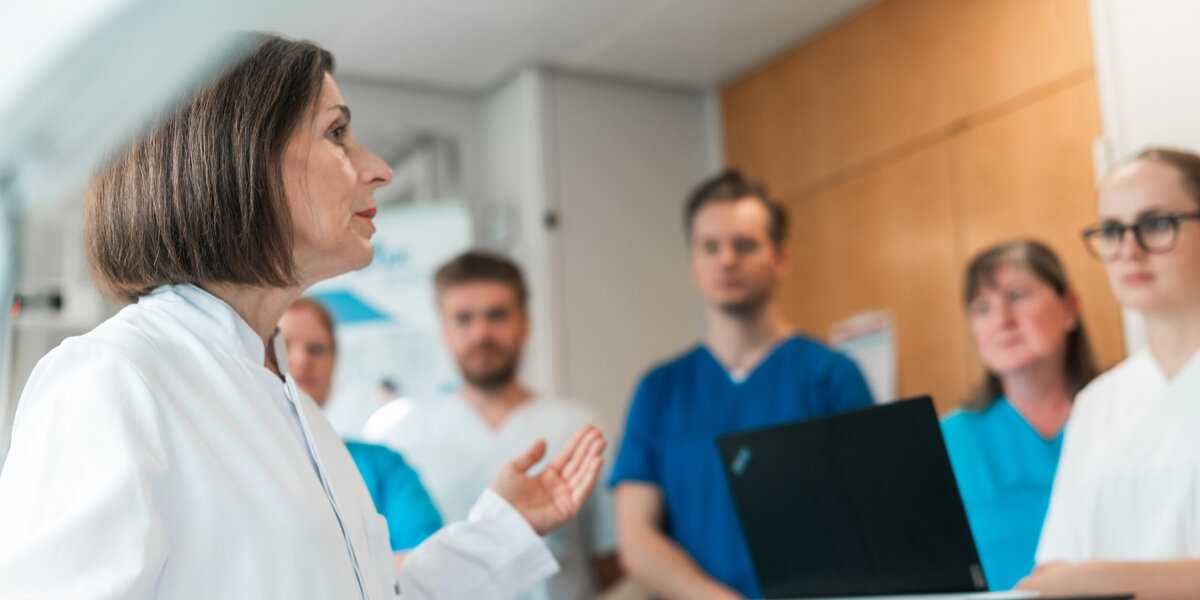 Eine Frau mit weißem Kittel steht vor einer Gruppe von jüngeren Personen; sie erklärt etwas und begleitet ihre Wort mit einer Geste