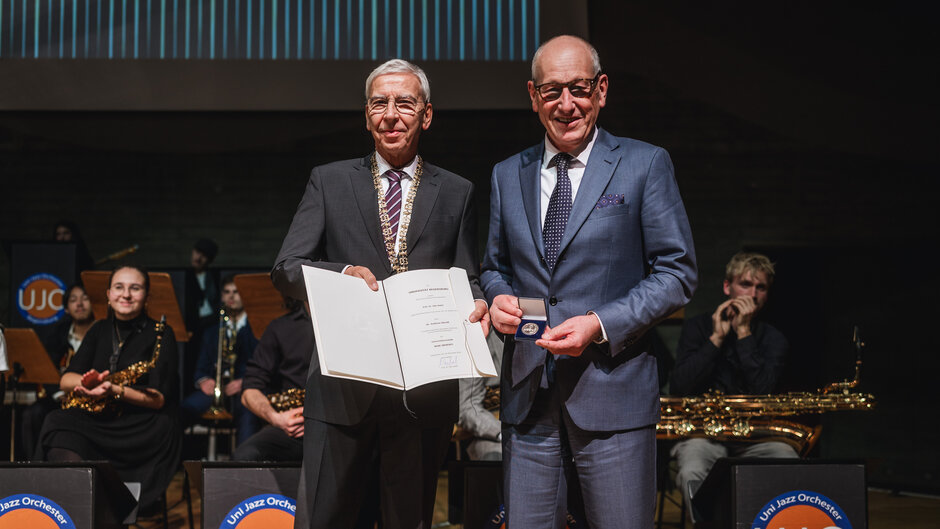Zwei Herren in dunklen Anzügen stehen auf der Bühne. Der links Stehende hält eine aufgeschlagene Urkundenmappe in der Hand und hält deren Inhalt in die Richtung des Betrachters. Der rechts stehende hält ein Futteral mit einer Medaille in der Hand und zeigt diese. 