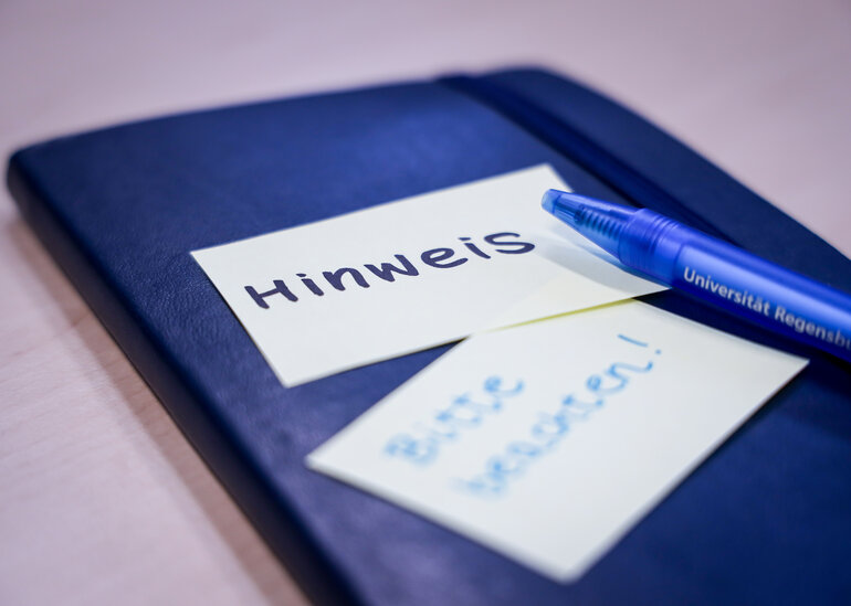 Notizbuch, auf dem ein Zettel klebt mit "Hinweis" und darunter ein Zettel mit "Bitte beachten". Darauf liegt ein blauer Kugelschreiber, auf dem Universität Regensburg steht.
