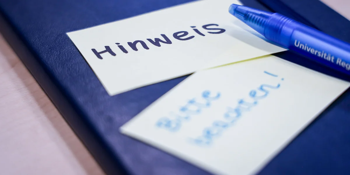 Notizbuch, auf dem ein Zettel klebt mit "Hinweis" und darunter ein Zettel mit "Bitte beachten". Darauf liegt ein blauer Kugelschreiber, auf dem Universität Regensburg steht.
