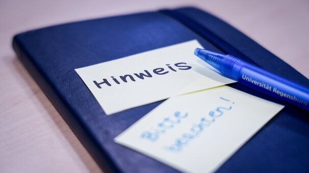 Notizbuch, auf dem ein Zettel klebt mit "Hinweis" und darunter ein Zettel mit "Bitte beachten". Darauf liegt ein blauer Kugelschreiber, auf dem Universität Regensburg steht.