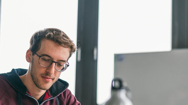 Drei junge Erwachsene sitzen an einem Tisch. Sie sind im Gespräch, eine Person macht Notizen, auf dem Tisch sind Tablets, Notizblöcke und eine Wasserflasche zu sehen.