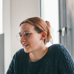 Drei junge Erwachsene sitzen an einem Tisch. Sie sind im Gespräch, eine Person macht Notizen, auf dem Tisch sind Tablets, Notizblöcke und eine Wasserflasche zu sehen.