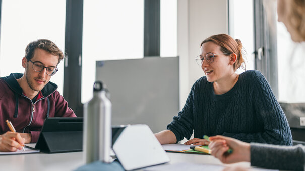 Drei junge Erwachsene sitzen an einem Tisch. Sie sind im Gespräch, eine Person macht Notizen, auf dem Tisch sind Tablets, Notizblöcke und eine Wasserflasche zu sehen.