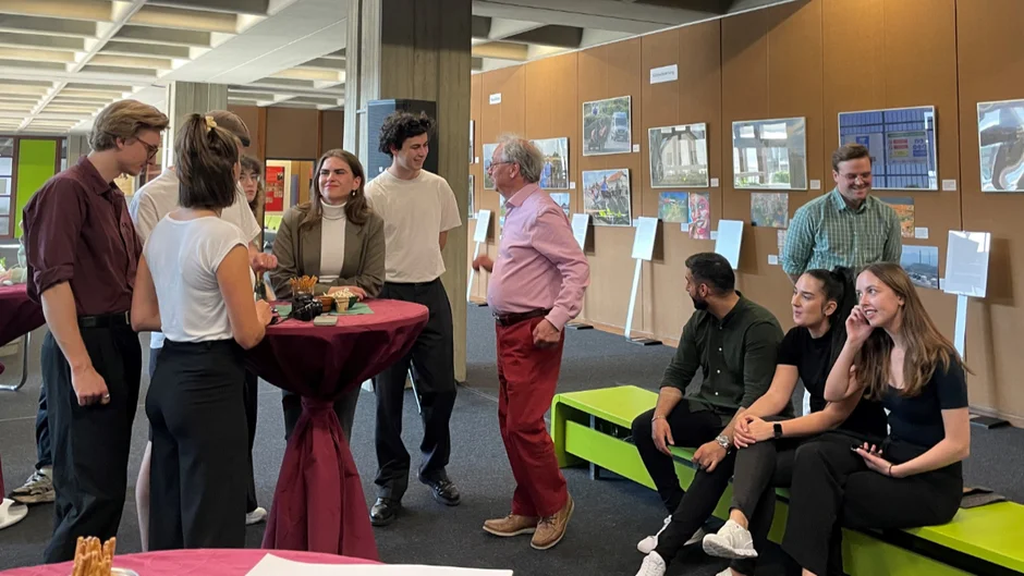 Mehrere Personen in einem Ausstellungsraum. Manche sind um einen Stehtisch gruppiert, andere sitzen auf einer grünen Bank; zwei weitere unterhalten sich. (öffnet Vergrößerung des Bildes)