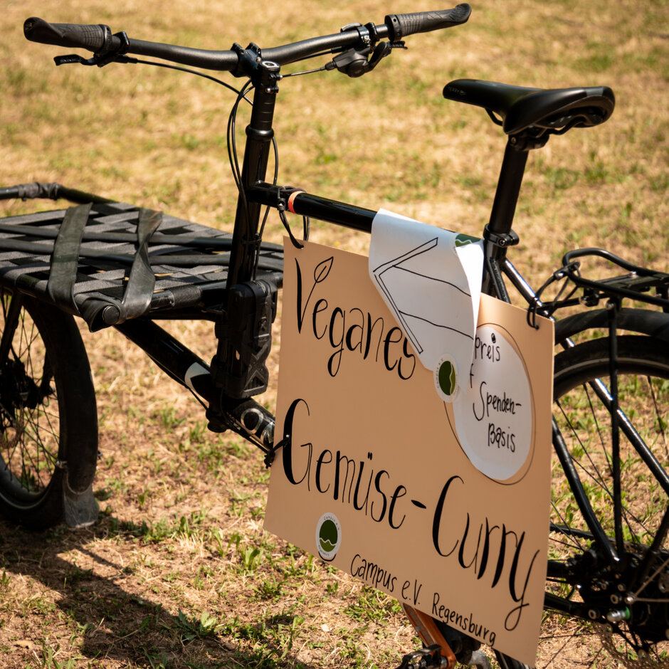 Auf einem Fahrrad ist ein Plakat montiert, auf dem steht: „Verganes Gemüse-Curry Preis auf Spenden-Basis Campus e.V. Regensburg". Darüber ist ein Pfeil angebracht, der nach links zeigt.