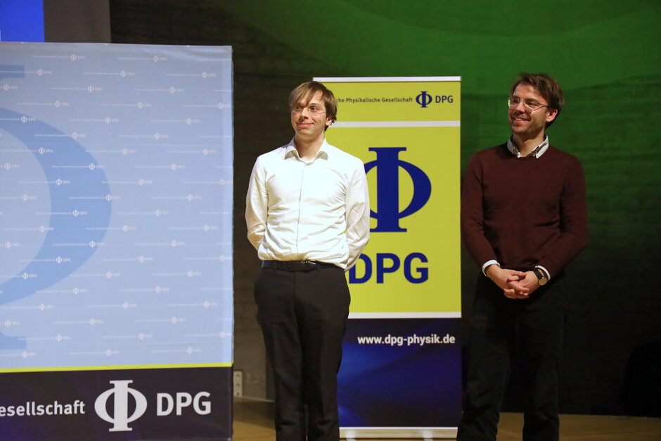 Zwei junge Männer stehen lächelnd neben Aufstellern mit DPG-Logo.