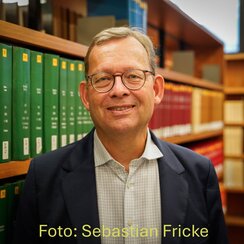 Portrait of a man in his sixties, wearing a suit, standing in front of a library shelf filled with books. Photo taken by Sebastian Fricke
