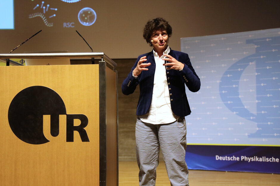 Eine Frau mit Kopfmikrophon steht gestikulierend neben einem mit UR-Logo geschmückten Rednerpult.