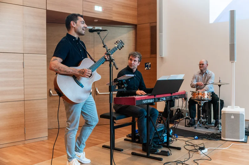 Auf dem Bild sind drei Männer zu sehen. Links singt ein Mann in ein Mikrofon und spielt dabei Gitarre, in der Mitte spielt ein Mann am Klavier, auf der rechten Seite spielt ein Mann Percussion. (öffnet Vergrößerung des Bildes)