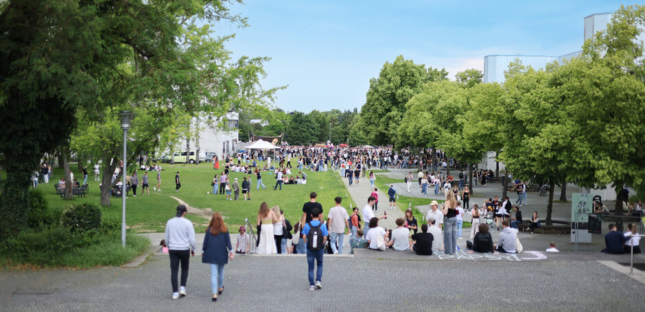 Blick über eine Wiese im Frühsommer, auf der sich viele junge Leute zum Feiern versammelt haben. Die Wiese ist umgeben von grünen Bäumen und Büschen, dahinter sind Hörsaalgebäude zu erkennen.