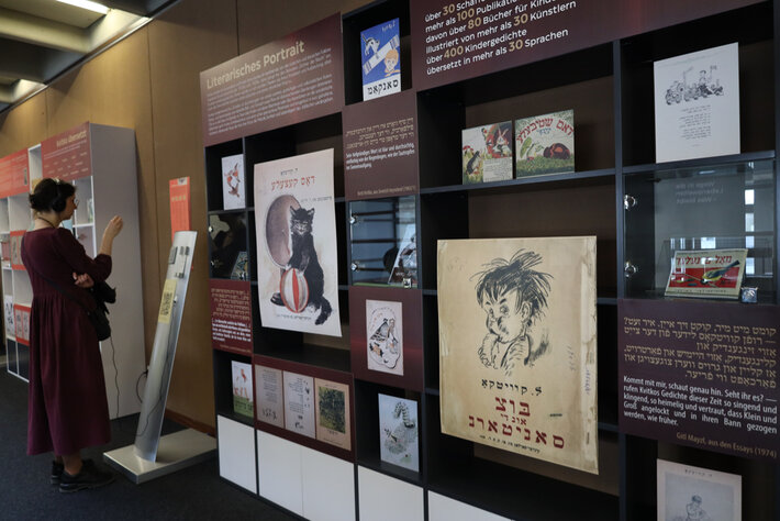 Blick auf eine Präsentationswand in einer Ausstellungsumgebung. Im Fokus ist die Zeichnung eines Kleinkindes, das sich etwas zu Essen in den Mund steckt. Darunter ist Text in hebräischen Schriftzeichen zu sehen. Einzelne Elemente der Wand sind mit Text in hebräischen Schriftzeichen bedruckt, darunter sind jeweils Übersetzungen in deutscher Sprache zu sehen. Die Wand ist überschrieben mit „Literarisches Portrait“. (öffnet Vergrößerung des Bildes)