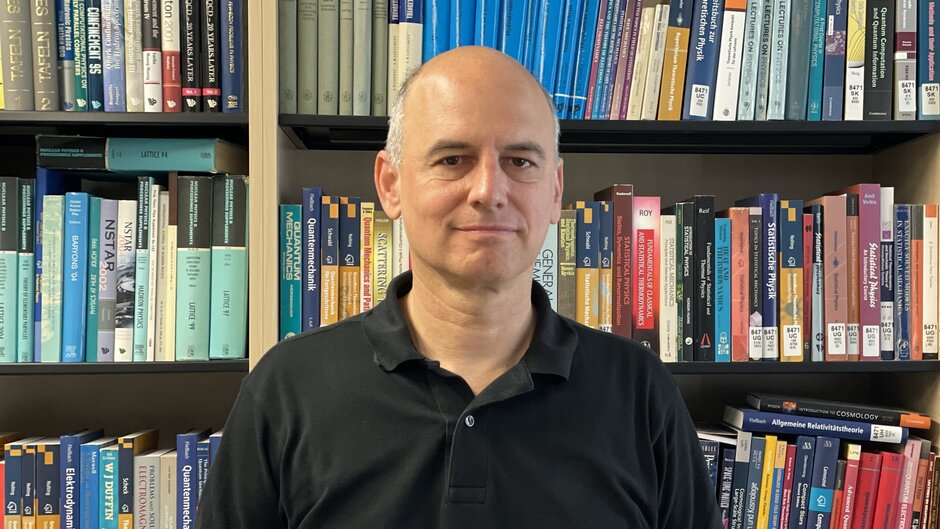 A man in a black polo shirt, standing in front of a bookshelf.