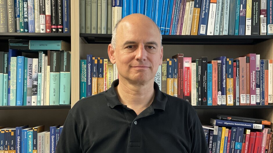 A man in a black polo shirt, standing in front of a bookshelf.