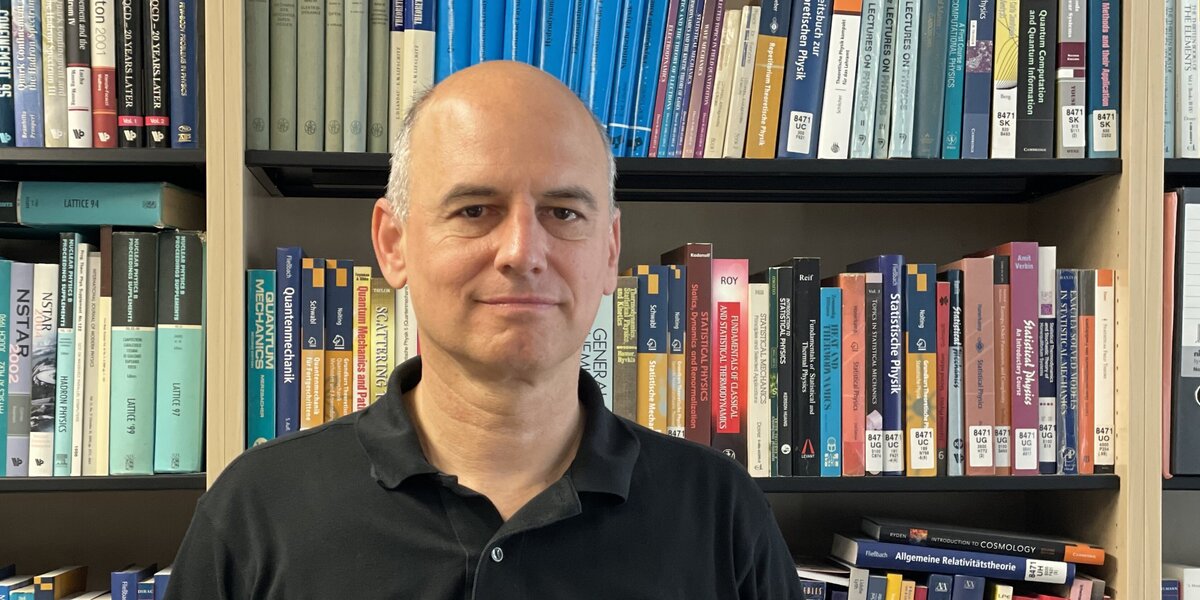 Bold man in black shirt standing in front of bookshelves.