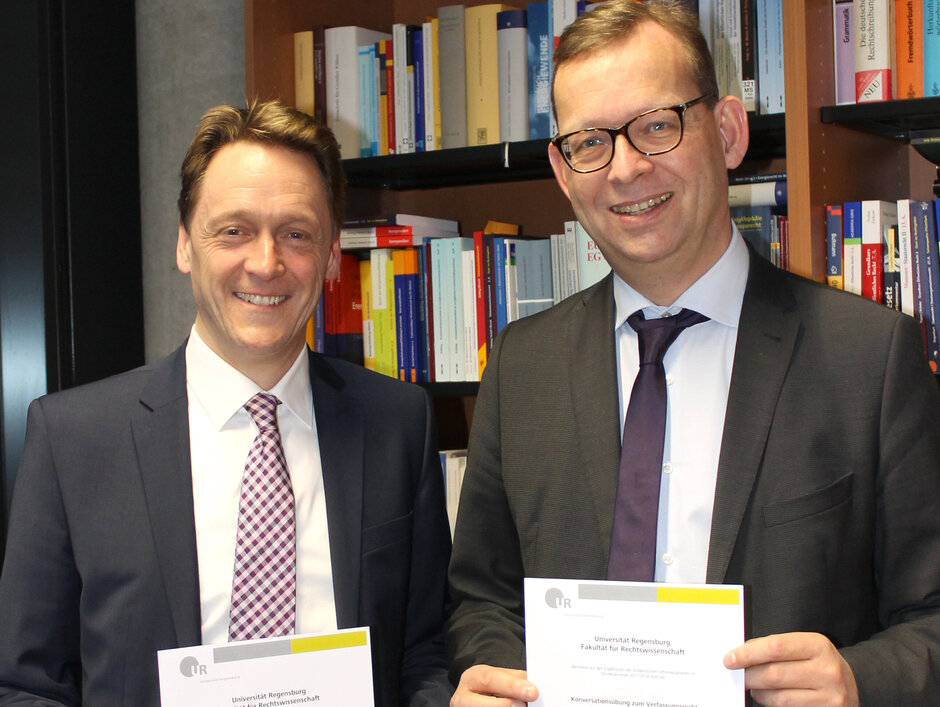 Prof. Kühling und Prof. Kingreen stehen lächelnd mit ihren Urkunden vor einem Bücherregal.