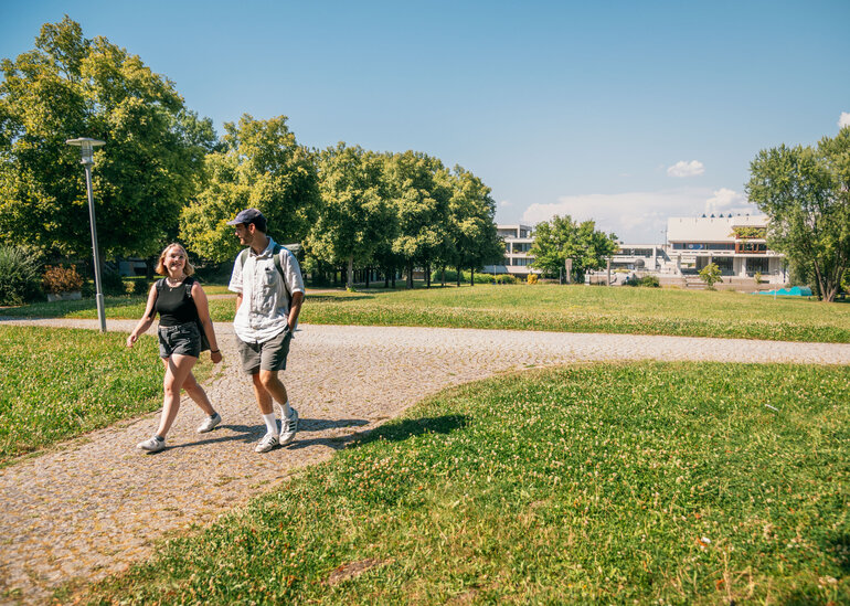 Zwei Studierende gehen im Sonnenschein, bei blauem Himmel und zwischen grünen Wiesen und Bäumen über den Campus der Universität Regensburg.