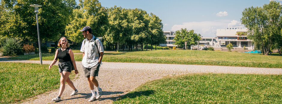 Zwei Studierende gehen im Sonnenschein, bei blauem Himmel und zwischen grünen Wiesen und Bäumen über den Campus der Universität Regensburg.