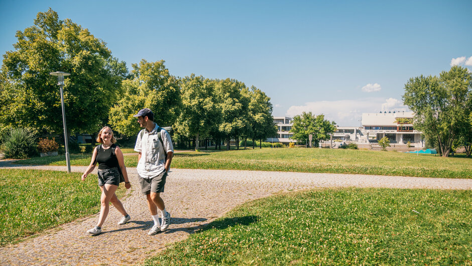 Zwei Studierende gehen im Sonnenschein, bei blauem Himmel und zwischen grünen Wiesen und Bäumen über den Campus der Universität Regensburg.