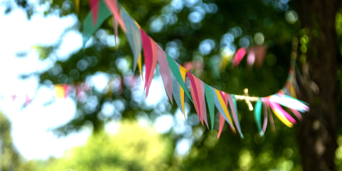 Eine Girlande bestehend aus bunten, dreieckigen Wimpeln flattert im Wind. Im Hintergrund sind grüne Bäume und blauer Himmel zu erahnen.