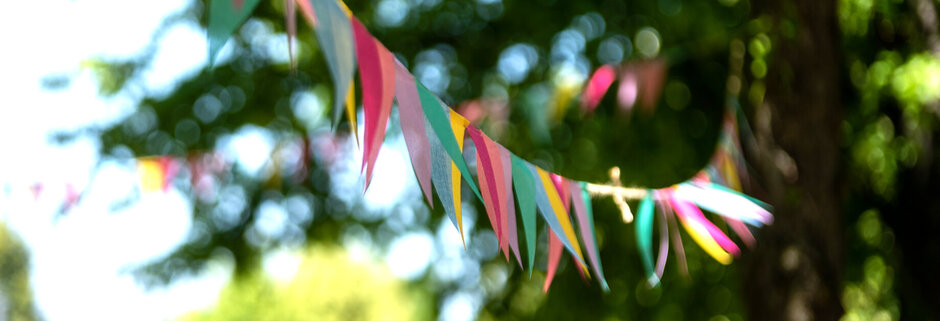 Eine Girlande bestehend aus bunten, dreieckigen Wimpeln flattert im Wind. Im Hintergrund sind grüne Bäume und blauer Himmel zu erahnen.