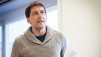 Ein Mann im grauen Strickpullover hält einen Papier in seiner linken Hand. Er scheint etwas zu erläutern.