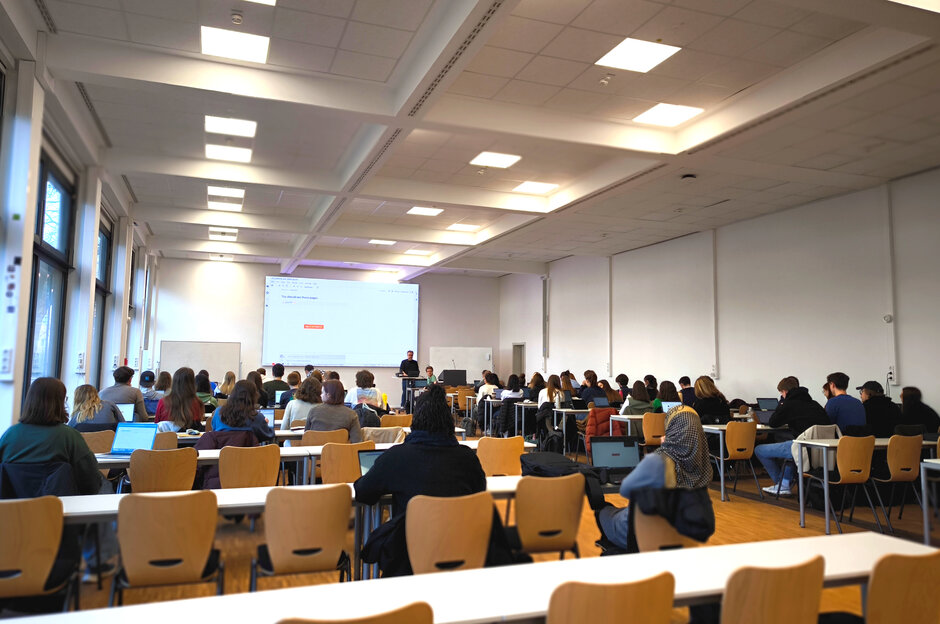 man sieht Studierende im Hörsaal von hinten, wie sie alle nach vorne zu einer Leinwand blicken.