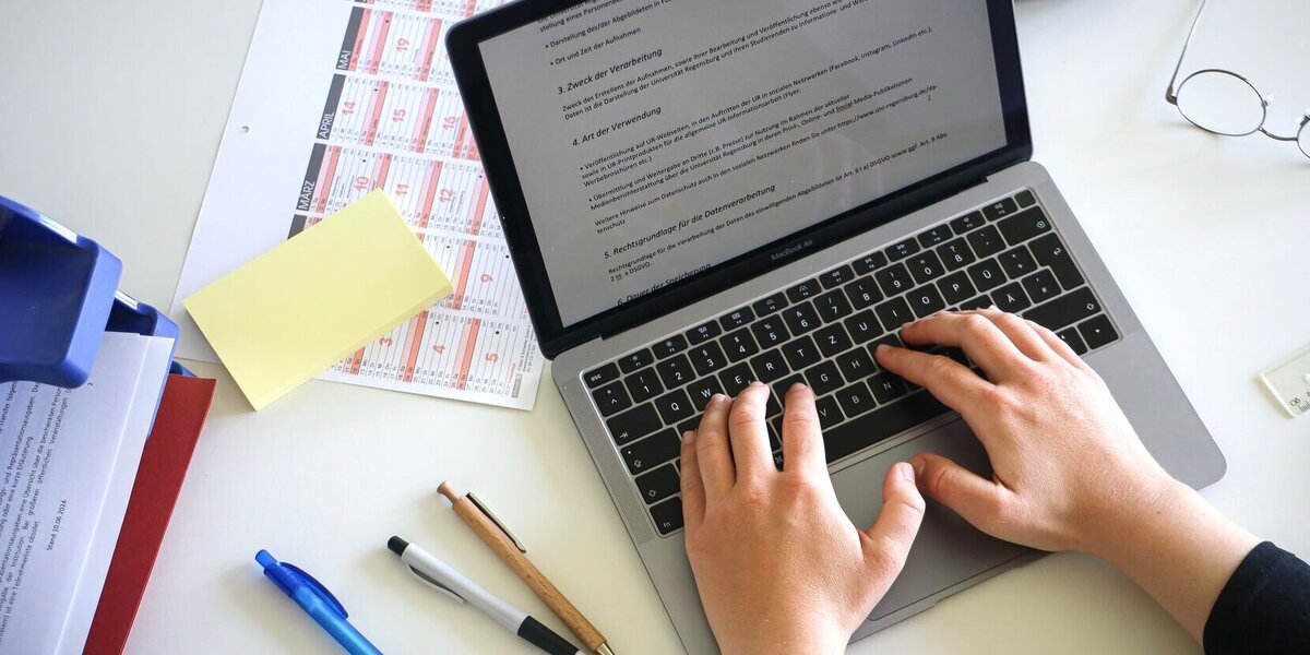 Person, die auf einem Laptop etwas schreibt. Daneben liegen Unterlagen und Stifte.