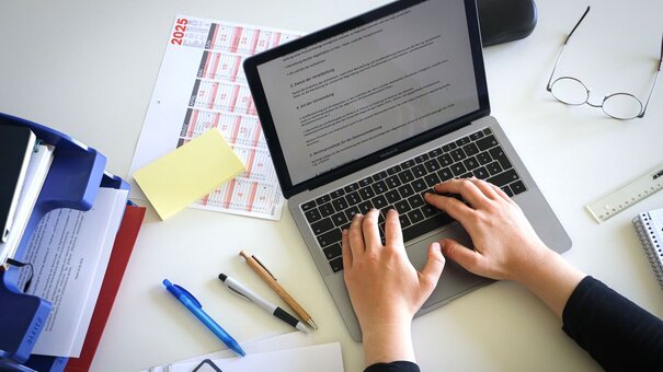 Person, die auf einem Laptop etwas schreibt. Daneben liegen Unterlagen und Stifte.