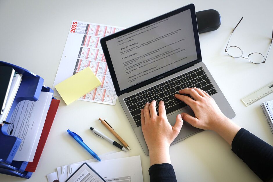 Person, die auf einem Laptop etwas schreibt. Daneben liegen Unterlagen und Stifte.