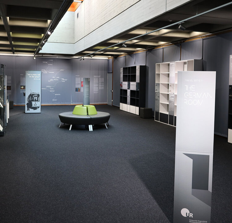 Ausstellungsraum schaufenster mit dunklem Teppichboden und Deckenbeleuchtung; links Glasvitrinen mit Dokumenten, rechts Regale und Informationstafeln. In der Mitte eine runde, grüne Sitzbank. Auf einem freistehenden Aufsteller steht „The German Room“, Tafeln zeigen Texte und Grafiken.